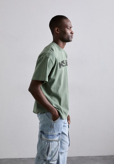 Grøn bomulds t-shirt med korte ærmer, prydet med et stort sort logo på fronten. Parret med lyseblå cargo jeans med flere lommer.