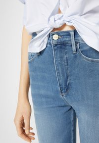 Jeans de mezclilla azul deslavada con cintura alta, dobladillo remangado y un botón de metal con el logo "FRAME", combinados con una camisa azul clara anudada.