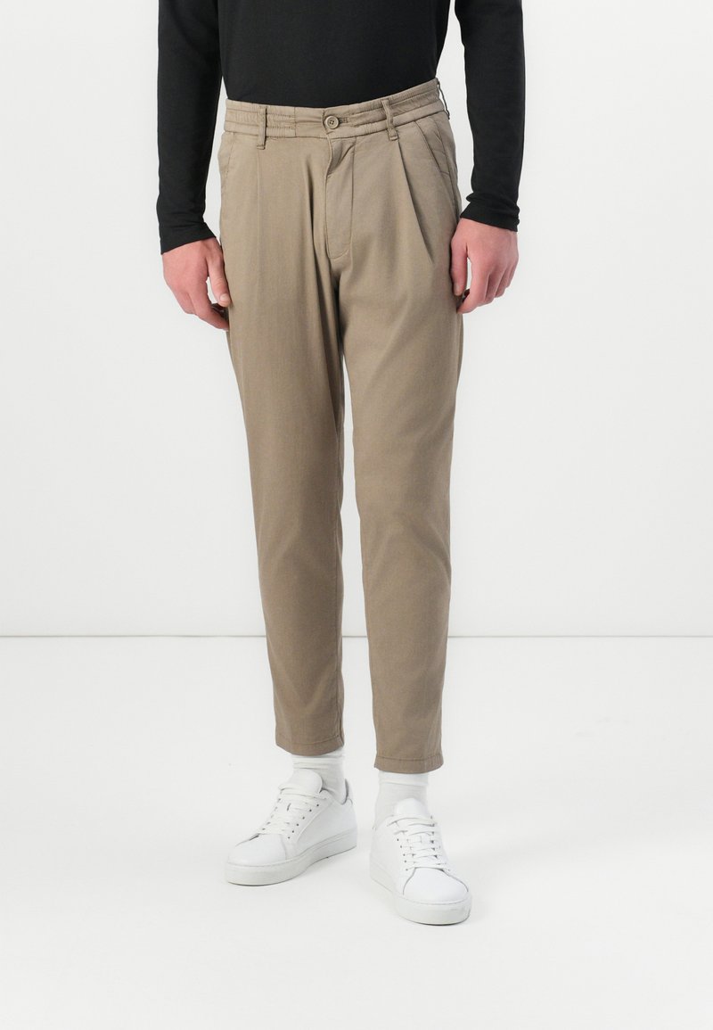 Beige Hosen mit Falten, aus leichtem Stoff. Kombiniert mit weißen Sneakers und knöchellangen Socken. Einfaches Design und lockere Passform.