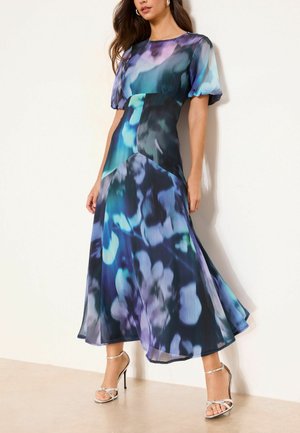 Femeie purtând o rochie midi vaporoasă cu modele abstracte în acuarelă albastre, verde albastrui și mov, cu mâneci bufante, asortată cu sandale argintii cu toc înalt.