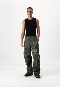 Svart ärmlös topp i kombination med löst sittande, kamouflagebyxor med cargo-stil. Har flera fickor och en texturerad yta. Bärs med mörka sneakers.