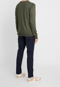 Henkilö pukeutuneena oliivinvihreään pitkähiharaiseen neuleeseen, tummansinisiiin housuihin, valkoisiin sukkiin ja beige-valkoisiin rentoihin tennareihin, seisoo yksinkertaista taustaa vasten.