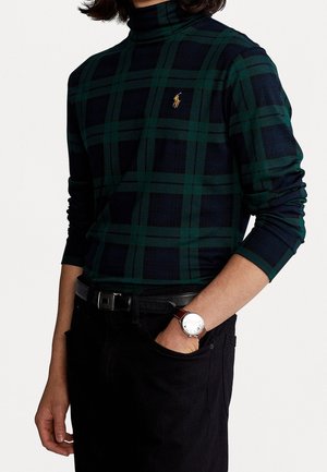 Persona vistiendo un suéter de cuello alto a cuadros verde oscuro y azul marino, pantalones negros, cinturón negro y reloj plateado con correa de cuero marrón, una mano en el bolsillo.