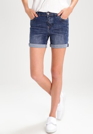 Shorts en denim avec un délavage bleu foncé, bords roulés et poches avant. Associés à des chaussures blanches à enfiler, portés avec un haut clair.