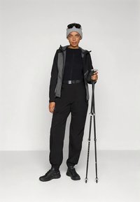 Crna vanjska jakna s sivom unutarnjom podstavom, odgovarajuće crne hlače i planinarske palice. Sivu kapu i sunčane naočale upotpunjuju odjeću.