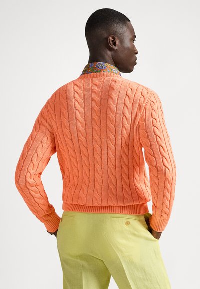 Orange cable-knit sweater with ribbed trim, worn over a patterned collared shirt. Paired with light green trousers featuring a back button detail.