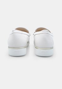 Un paio di scarpe bianche slip-on con retro texturizzato e suole bianche basse, viste da dietro su uno sfondo semplice.
