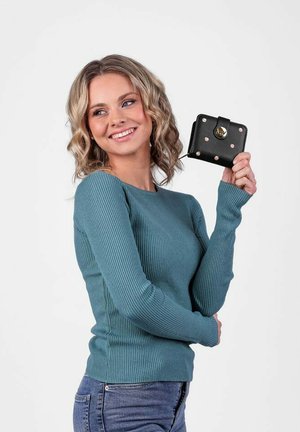 Teal ribbed sweater, long sleeves, fitted silhouette. Model holding a black wallet with round gold hardware and pink studs, against a white background.