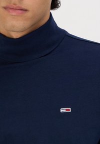 Dunkelblauer Rollkragenpullover aus weichem Stoff. Verfügt über ein kleines, gesticktes Logo mit roten, weißen und blauen Akzenten auf der Brust.