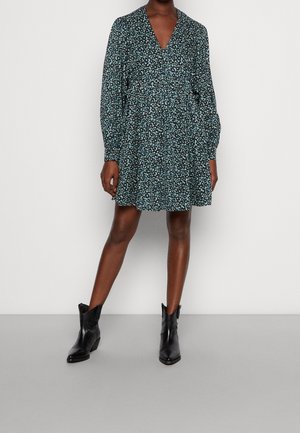 Femme portant une robe noire et turquoise à manches longues, longueur au-dessus du genou, avec des bottines noires, debout devant un fond blanc uni.