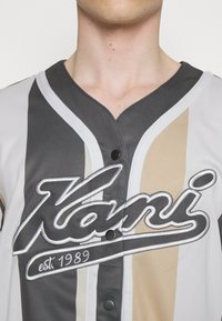 Grå, beige og hvit baseballtrøye med knapper og V-hals, med brodert "Kani" logo og "est. 1989" detalj. Glatt tekstur.