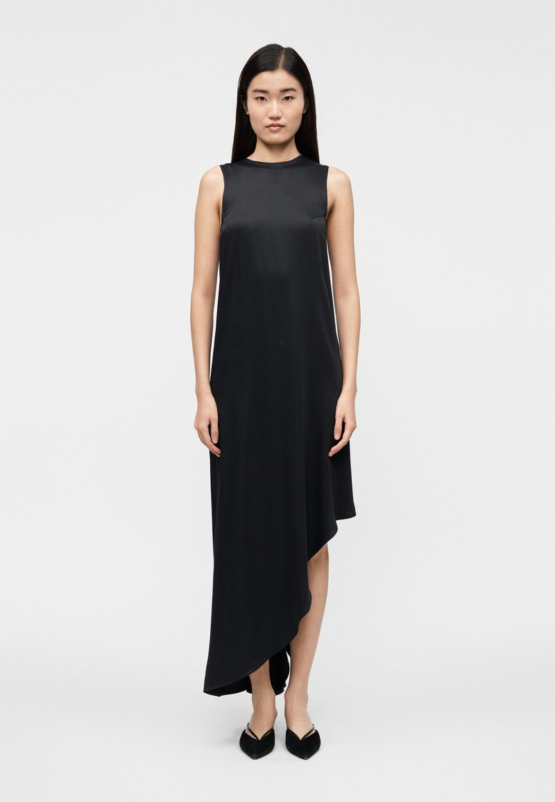 Vestido preto sem mangas com decote alto, bainha assimétrica e textura suave, combinado com sapatos planos pontiagudos.