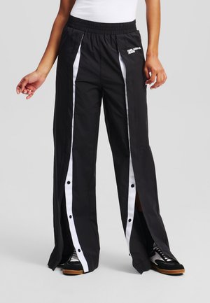 Pantalones deportivos negros con franjas blancas a los lados, cintura elástica y aberturas con botones a presión a lo largo de los costados. Fabricados con un tejido suave.