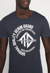 Navy blue cotton t-shirt featuring a large white graphic with the text "Quality Denim Brand" and a diamond logo, round neckline, and short sleeves.