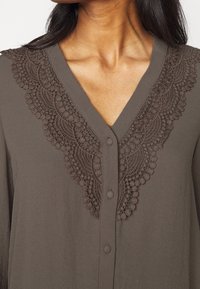 T-shirt a V in tessuto marrone scuro con intricate ricami in pizzo sul colletto. Presenta chiusura con bottoni e una consistenza morbida e liscia.