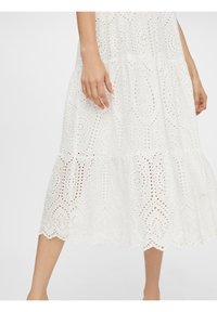 Witte jurk van ingewikkelde eyelet stof, met een gelaagd ontwerp, een gekartelde zoom en gedetailleerde patronen doorheen.