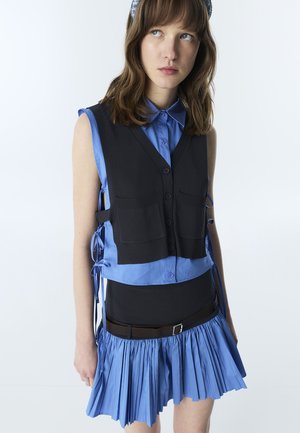 Giovane donna che indossa una gonna blu plissettata e una camicia, abbinata a un gilet nero senza maniche con bottoni, legato ai lati, che guarda verso sinistra.