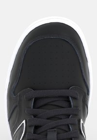 Close-up of the black sneaker toe with perforations, black laces, and white rubber sole visible at the edges.