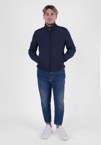 Giacca zip-up blu navy con colletto alto, texture morbida e tasche laterali, abbinata a jeans blu con risvolto e sneakers bianche.