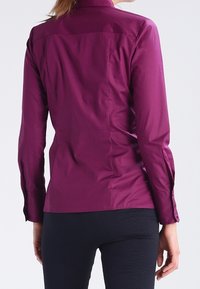 Camicia aderente a maniche lunghe di un profondo viola con una texture liscia, cucitura a yoke sul retro e polsini con bottoni. Abbinata a pantaloni neri testurizzati.