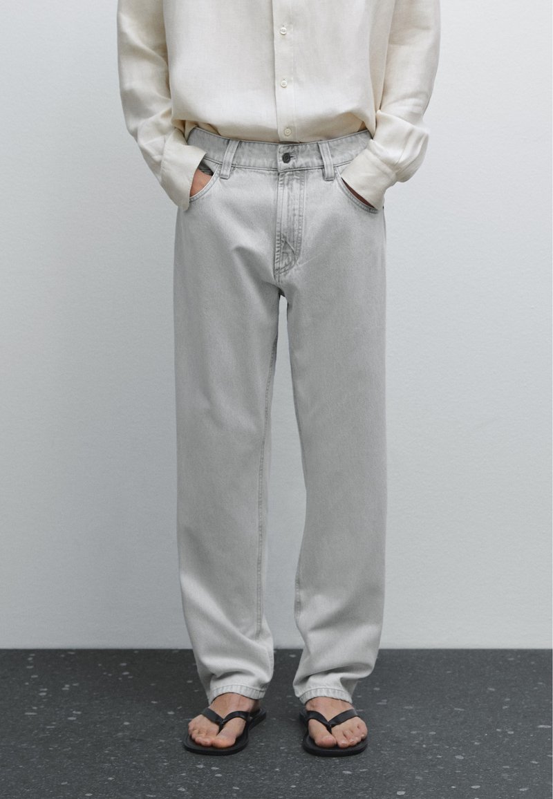 Lysegrå denim jeans med en afslappet pasform, der har et klassisk fem-lomme-design, lukning med topknap og en slidt, blød tekstur. Båret med sorte flip-flops.