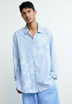 Noor mees kannab lahtist helesinist triibulise ja lillemustriga õmblustega särki, mida täiendavad sobivad sinised triibulised püksid.