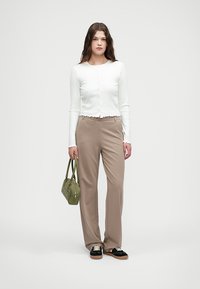 Biała bluzka z długim rękawem i zapięciem na guziki, z falbaniastym wykończeniem, w połączeniu z beżowymi spodniami z szerokimi nogawkami. Modelka trzyma zieloną torebkę z ćwiekami.