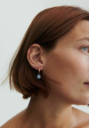 Sieviete ar īsiem brūniem matiem, valkājot zelta riņķa auskaru ar mazu apaļu zilu pērlīti, skatās pa labi uz vienkārša gaiša fona.