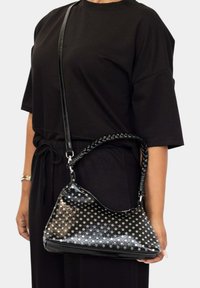 Bolso de mano negro con un patrón de lunares, material de cuero y una correa de hombro trenzada, sostenido de lado por una persona.