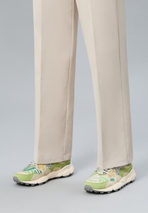 Personne portant un pantalon beige à jambes droites et des baskets à motifs verts, blancs et beiges sur un fond gris.