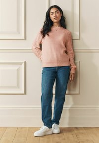 Gaiši rozā džemperis ar teksturētiem, mežģīņu piedurknēm, kopā ar zilām džinsu biksēm un baltām sporta kurpēm. Modelis stāv pie neitrālas sienas.