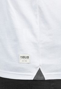 Camiseta blanca de manga corta con una etiqueta de marca rectangular. Presenta un dobladillo profundo con tela gris en contraste en las ventilaciones laterales, textura suave.