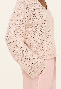 Helles rosa Strickpullover mit V-Ausschnitt, der über offene Muster, strukturierte Ärmel und einen gewellten Saum verfügt. Kombiniert mit weichen rosa Hosen.