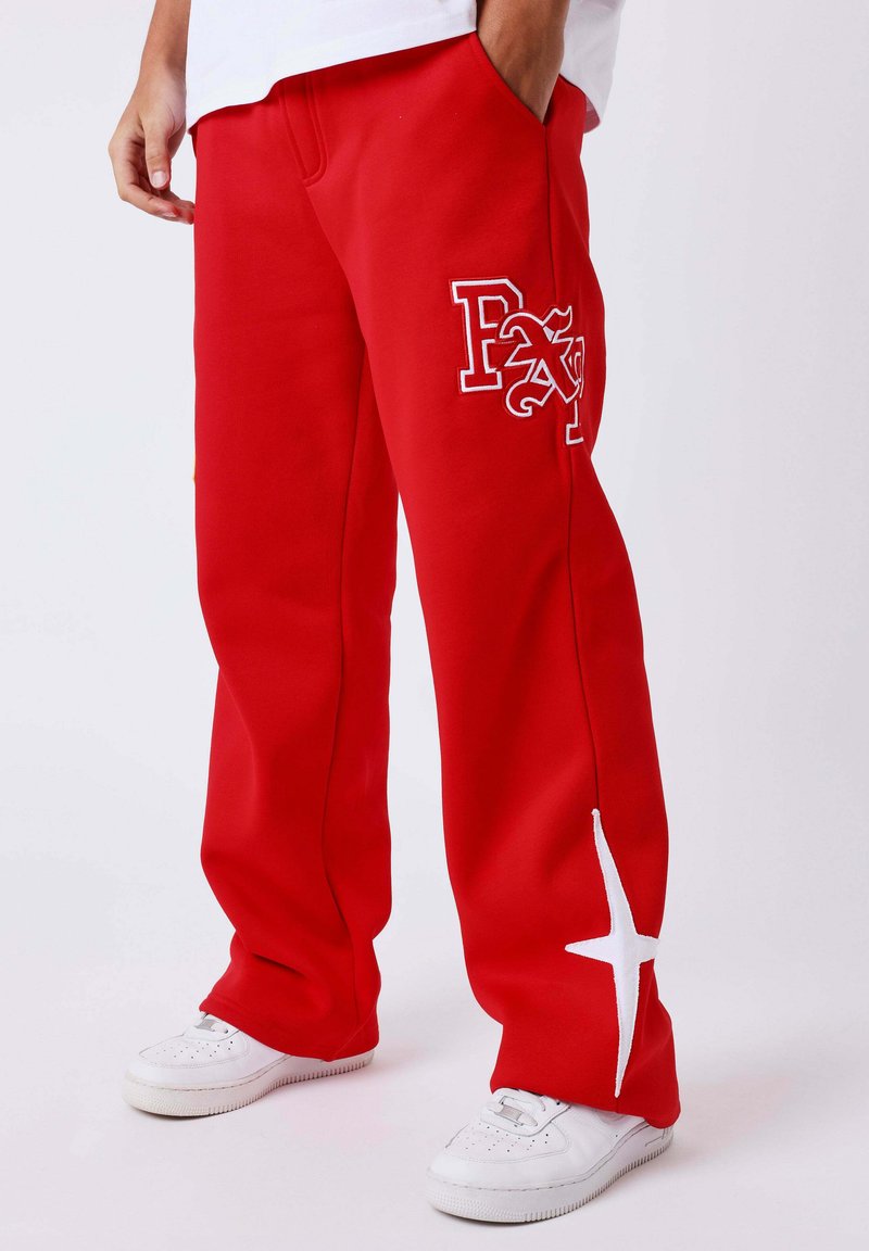 Personne portant un pantalon rouge avec les lettres "PXL" brodées en blanc sur la cuisse et un motif d'étoile blanche près de la cheville, associé à des baskets blanches.