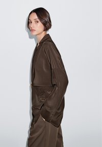 Camicia oversize in satin marrone con colletto a punta, maniche lunghe e tasca laterale, caratterizzata da una texture liscia e da un design minimalista.