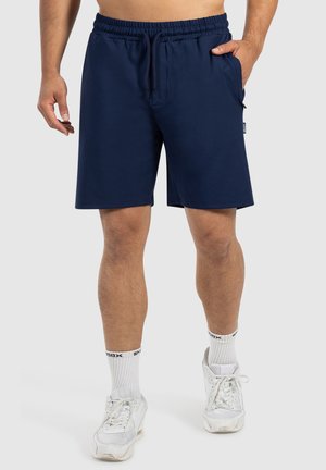 Marineblauwe sportshorts van zachte stof, met een elastische tailleband, trekkoord en zijzakken. Draagt met witte sneakers en sokken.