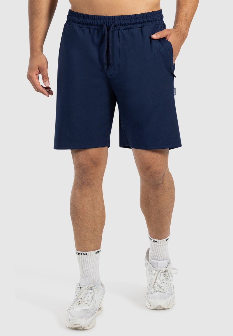 Marineblauwe sportshorts van zachte stof, met een elastische tailleband, trekkoord en zijzakken. Draagt met witte sneakers en sokken.