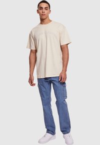 Camiseta de algodón beige con el texto "Urban Classics", combinada con pantalones cargo de mezclilla azules y zapatillas blancas, que presentan un corte relajado y mangas cortas.