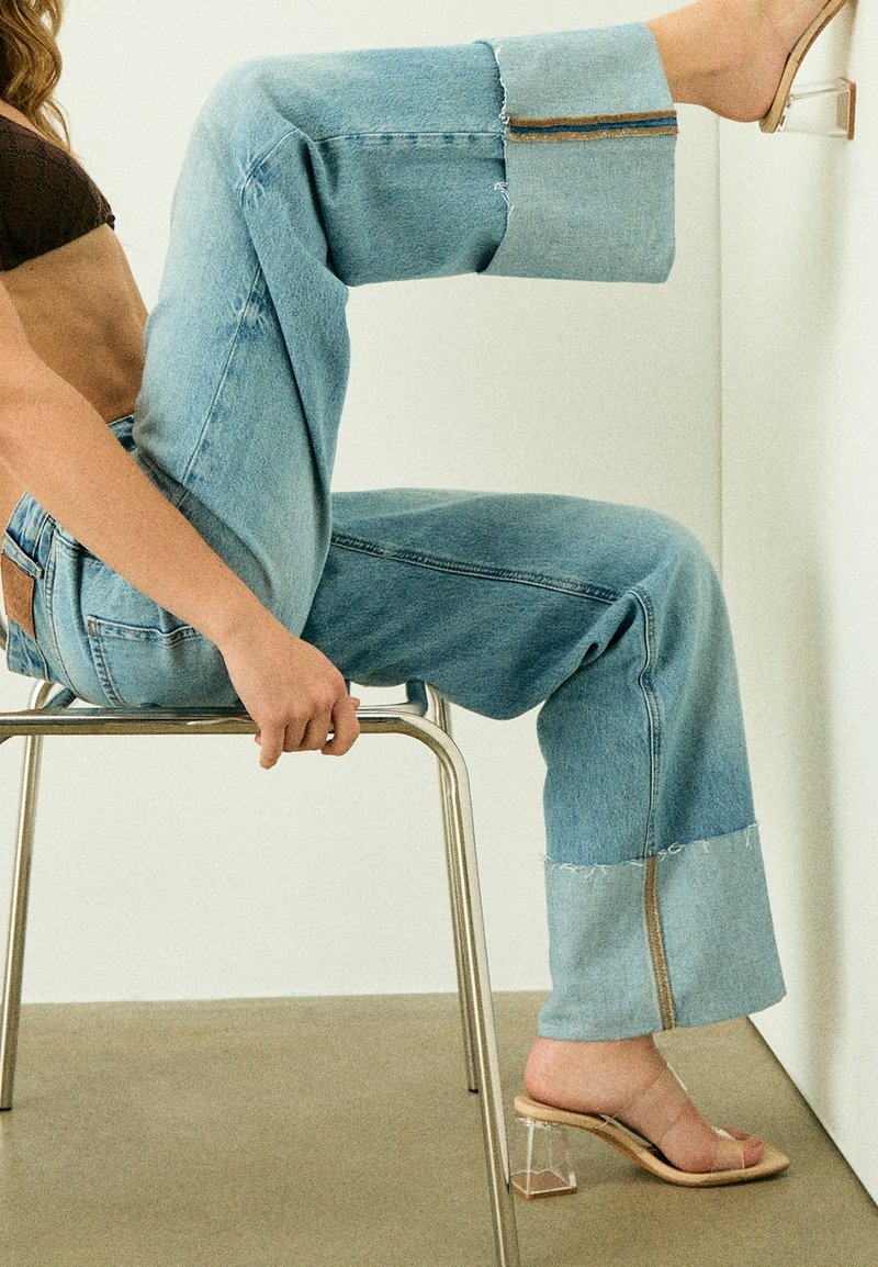 Femme en jeans bleus et sandales à talons transparents assise de côté sur une chaise, une jambe pliée avec le pied reposant contre un mur blanc.