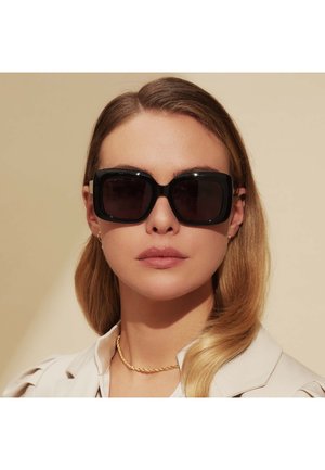 Schwarze übergroße Sonnenbrille mit quadratischen Gläsern, glänzender Oberfläche, mit beigen Seitenakzenten, getragen zu einem hellen Kragenoberteil.