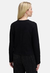 Blondynka zwrócona w prawo, ubrana w strukturalny czarny długi rękaw, a także błyszczące czarne spodnie, stojąca na tle jasnym.