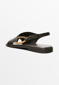 Schwarze Ledersandale mit flacher Sohle, offenem Fersenbereich und einem auffälligen goldenen Metallelement. Verfügt über ein minimalistisches Design und eine glatte Textur.
