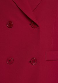 Dubbelzijdig jasje van dieprode stof, met glanzende rode knopen, scherpe revers en een zijzakdetail. Zachte textuur.