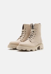 Botas beige con cordones, suelas gruesas y cremalleras laterales, con adornos plateados decorativos en el interior de la bota derecha.