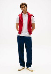 Hvid polo-shirt, rød hættetrøje draperet over skuldrene, blå bukser, hvide sneakers med beige såler. Enkel design og rene linjer.