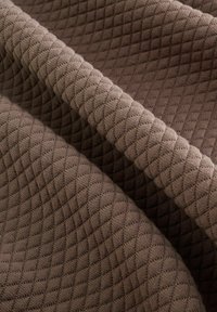 Tessuto trapuntato marrone con motivo a diamante, superficie strutturata e pieghe che creano profondità. Il materiale sembra morbido e resistente.