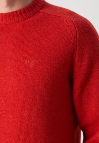 Pull col rond rouge en tricot avec un logo Barbour ton sur ton subtilement brodé sur la poitrine, porté par une personne montrée du cou au haut du torse.