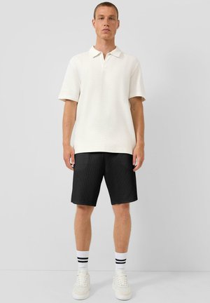 Junger Mann steht und trägt ein weißes kurzärmeliges Polohemd, schwarze knielange Shorts, weiße Socken mit schwarzen Streifen und weiße Turnschuhe.