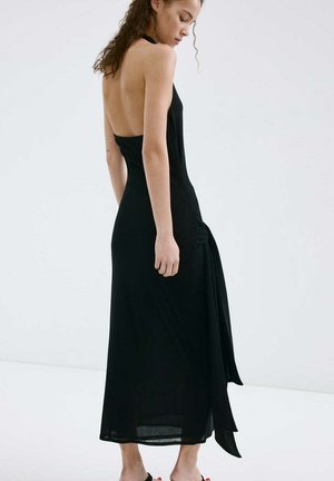 Mujer con pelo rizado que lleva un vestido midi negro sin mangas y sin espalda, y tacones negros con tiras, de pie frente a un fondo blanco liso.