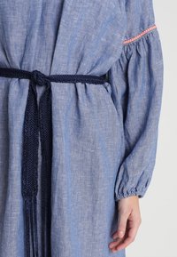 Vaaleansininen pellavamekko, jossa on tummansininen punottu vyö, puffihihat ja vaaleanpunaisia korostustikkoja hihojen kohdalla. Suora pinta.